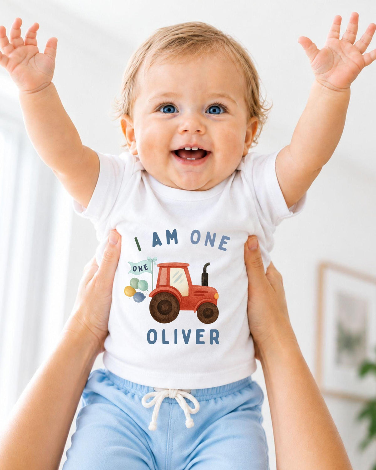 A smiling baby wearing a white shirt with a tractor graphic and the text "I am one Oliver" is being held up by someone's arms.