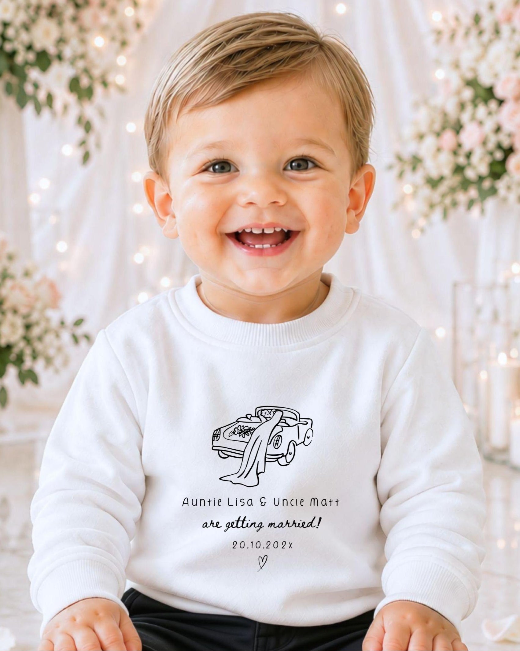 A smiling baby boy wearing a white sweater with a graphic design and text on it, sitting in front of a floral background.