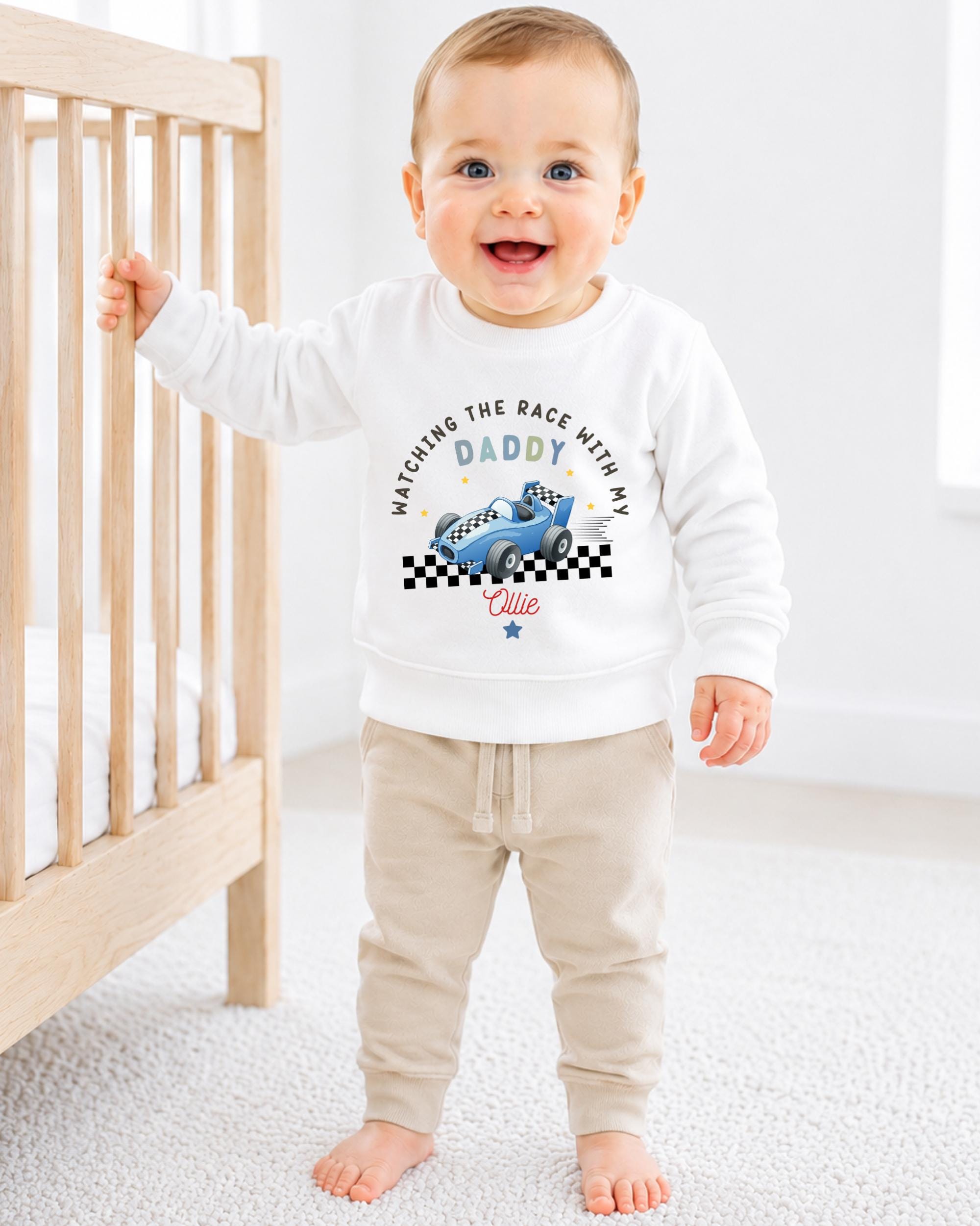 A smiling baby boy wearing a white sweatshirt with a racing car design and the text "Watching the race with my Daddy with my name is Ellie" is standing in front of a wooden crib.