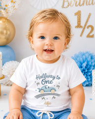 A smiling baby boy wearing a white t-shirt with a graphic of a car and the text "Halfway to One" is sitting in front of a birthday party backdrop.