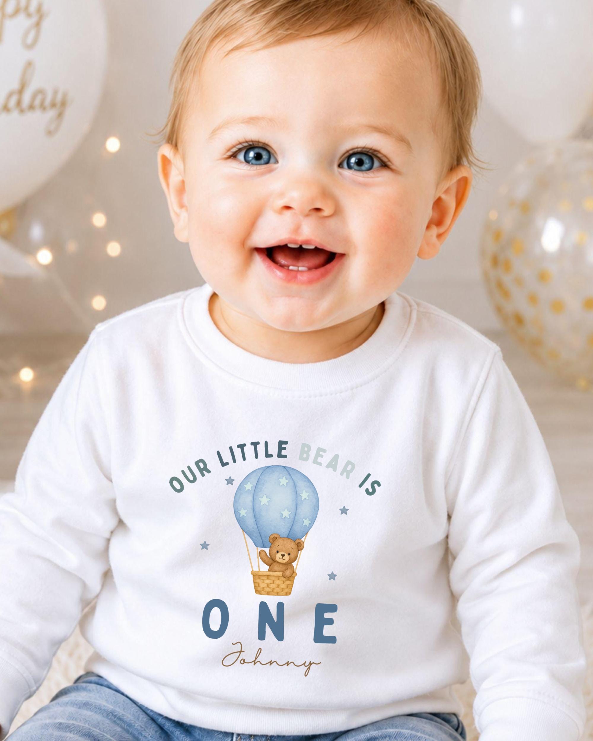 A smiling baby boy wearing a white shirt with a teddy bear and a hot air balloon graphic, celebrating his first birthday.