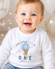 A smiling baby boy wearing a white shirt with a teddy bear and a hot air balloon graphic, celebrating his first birthday.