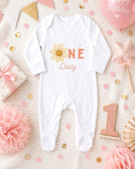 A white onesie with the text "Daisy" printed on it, surrounded by pink and gold decorations, including a pink star, confetti, and a pink balloon with the text "Happy Birthday".