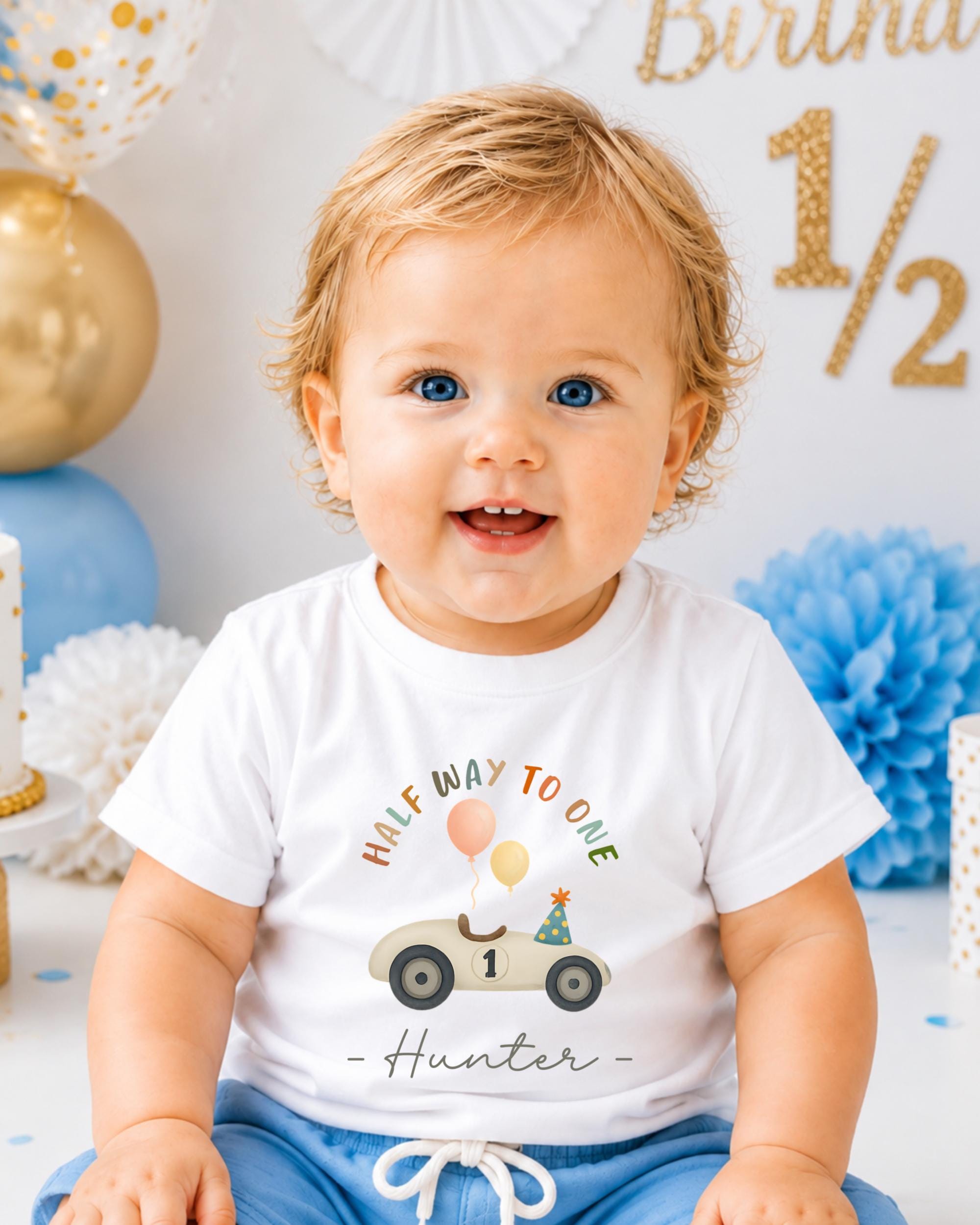 A smiling baby boy wearing a white t-shirt with a birthday-themed graphic and the text "Half way to one Hunter" is sitting in front of a birthday party backdrop.