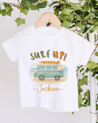 A white t-shirt with a graphic design of a van and the text "Surf up! Jackson" printed on it, hanging on a wooden hanger.