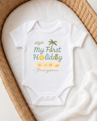 A white onesie with a printed design of a palm tree, airplane, and beach, along with the text "My First Holiday - Grayson" printed on it.