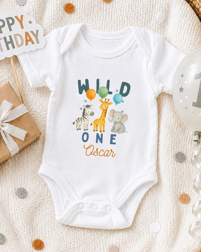 A white bodysuit with a design of wild animals, including a giraffe, zebra, and elephant, along with the text "Wild One Oscar" printed on it. The bodysuit is placed on a white blanket, surrounded by various party decorations such as balloons and a "Happy Birthday" sign.