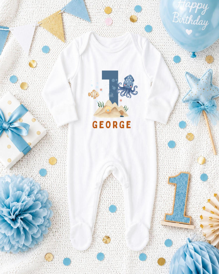 A white bodysuit with the number 1 and an octopus design, surrounded by birthday decorations such as a blue balloon, blue and gold confetti, and a blue and gold star.