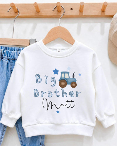 A white sweatshirt with a tractor design and the text "Big Brother Matt" printed on it, hanging on a wooden hanger.