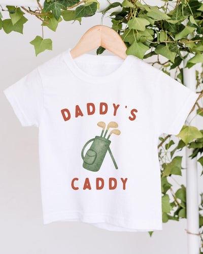 A white t-shirt with a graphic design of a golf bag and the text "DADDY'S CADDY" printed on it, hanging on a wooden hanger.