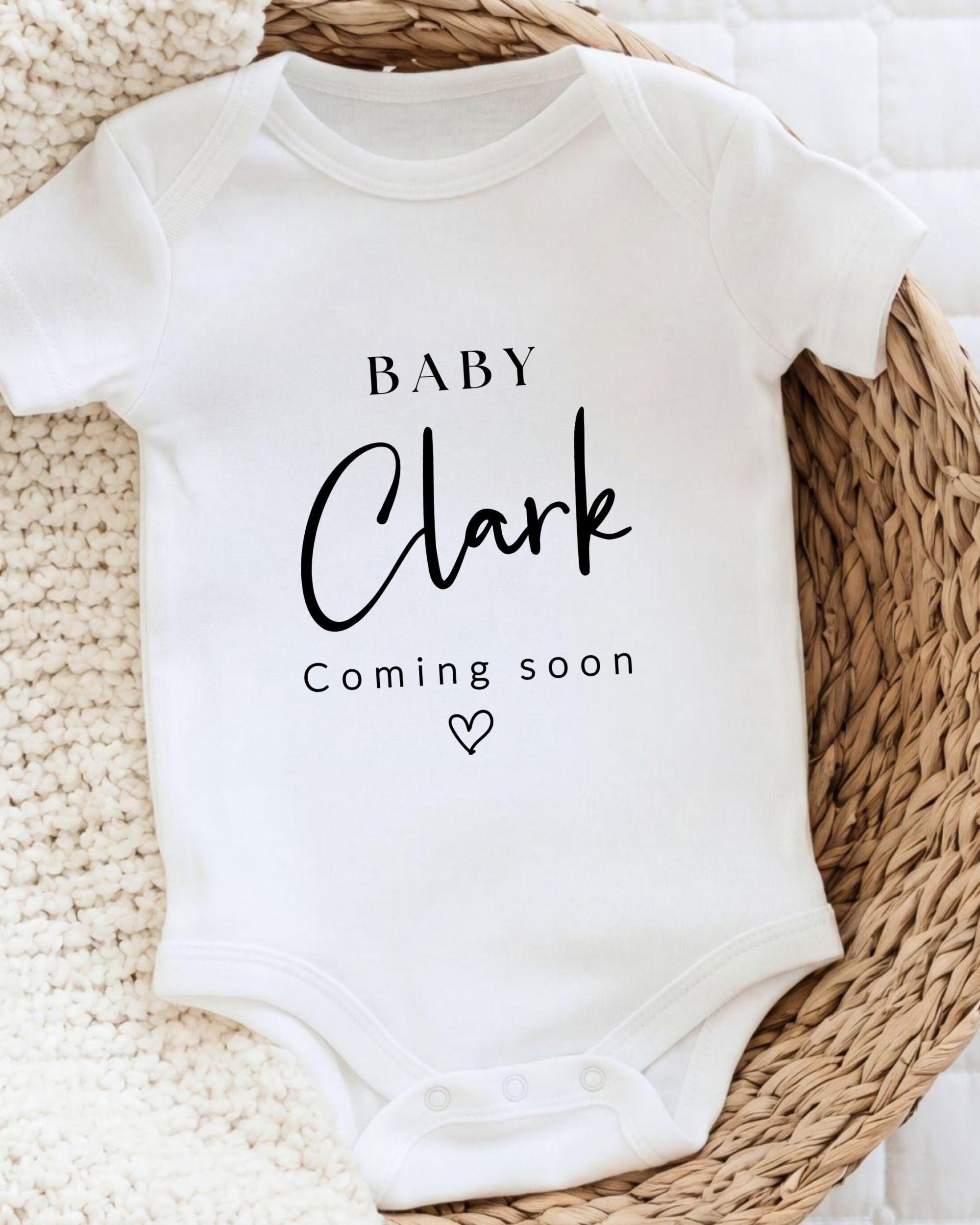 A white baby onesie with the text "BABY Clark Coming soon" printed on it, placed on a woven basket.