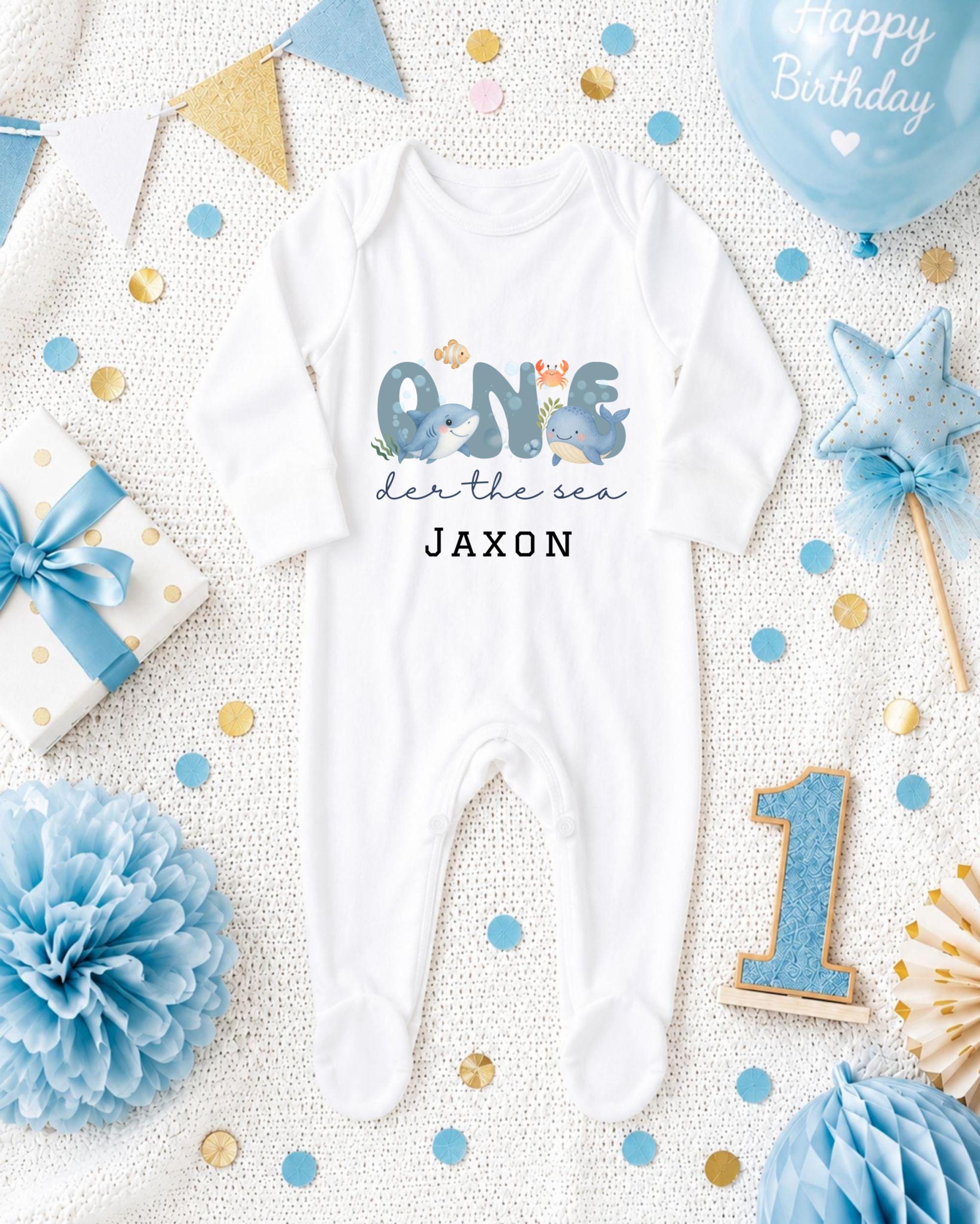 A white onesie with the text "ONE den the sea JAXON" printed on it, surrounded by various baby-themed items such as a blue balloon, a blue and gold star, and blue and gold confetti.