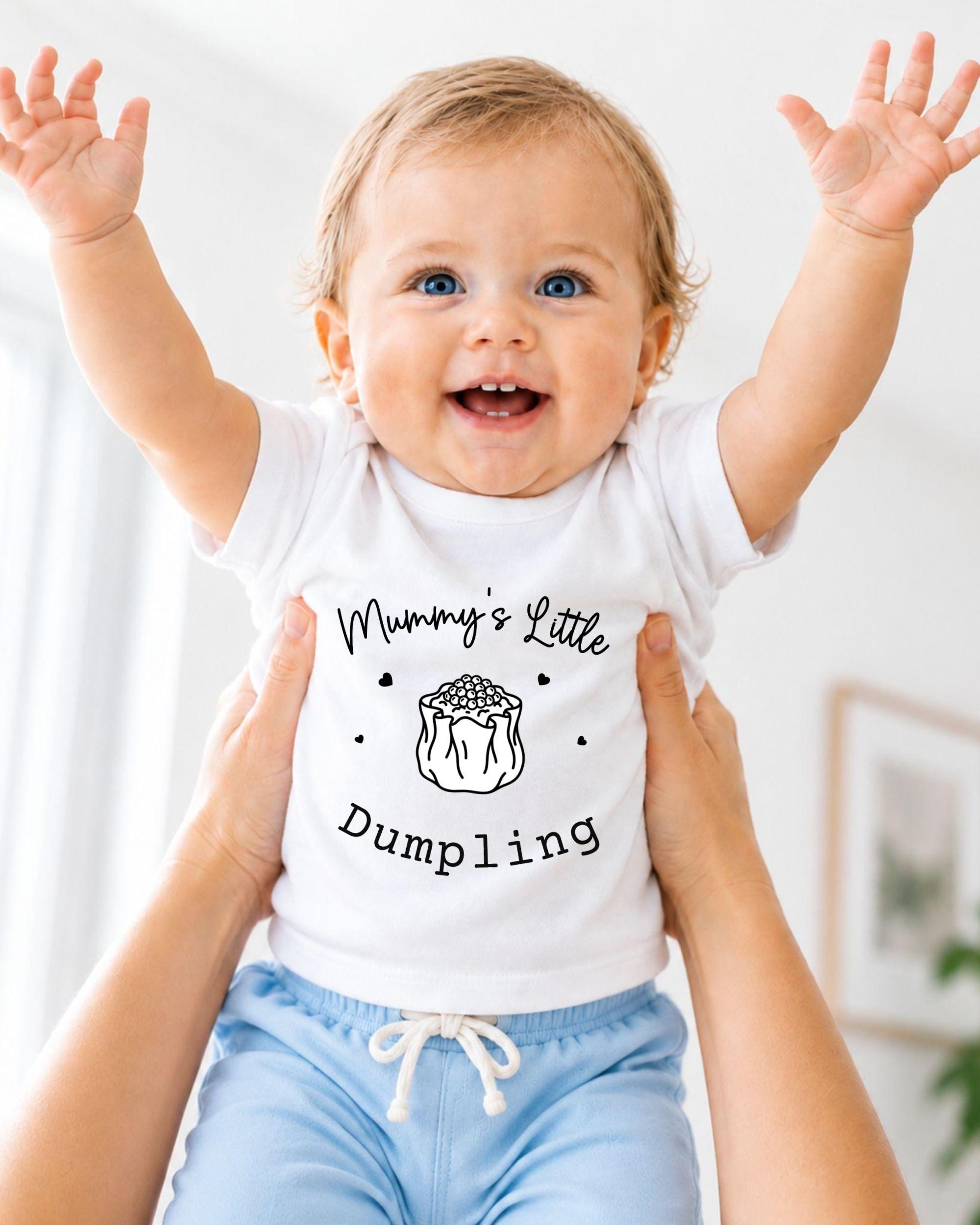 A smiling baby wearing a white t-shirt with the text "Mummy's Little Dumping" is being held up by someone's arms.