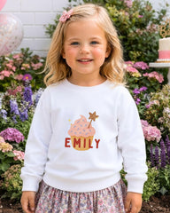 A young girl with blonde hair is wearing a white sweater with a cupcake design and the name "EMILY" printed on it, standing in front of a garden with colorful flowers.