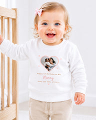 A smiling baby girl wearing a white sweatshirt with a heart-shaped photo and text on it.