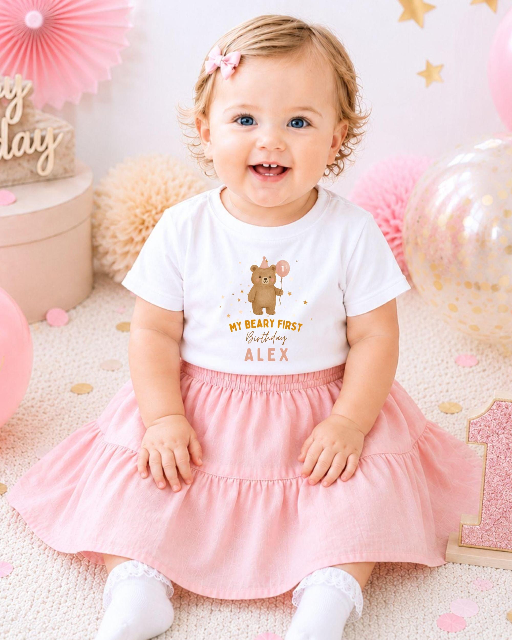 A smiling baby girl wearing a white t-shirt with a cartoon bear and the text "My heart first birthday Alex" sits on a pink and white background with pink and gold decorations.
