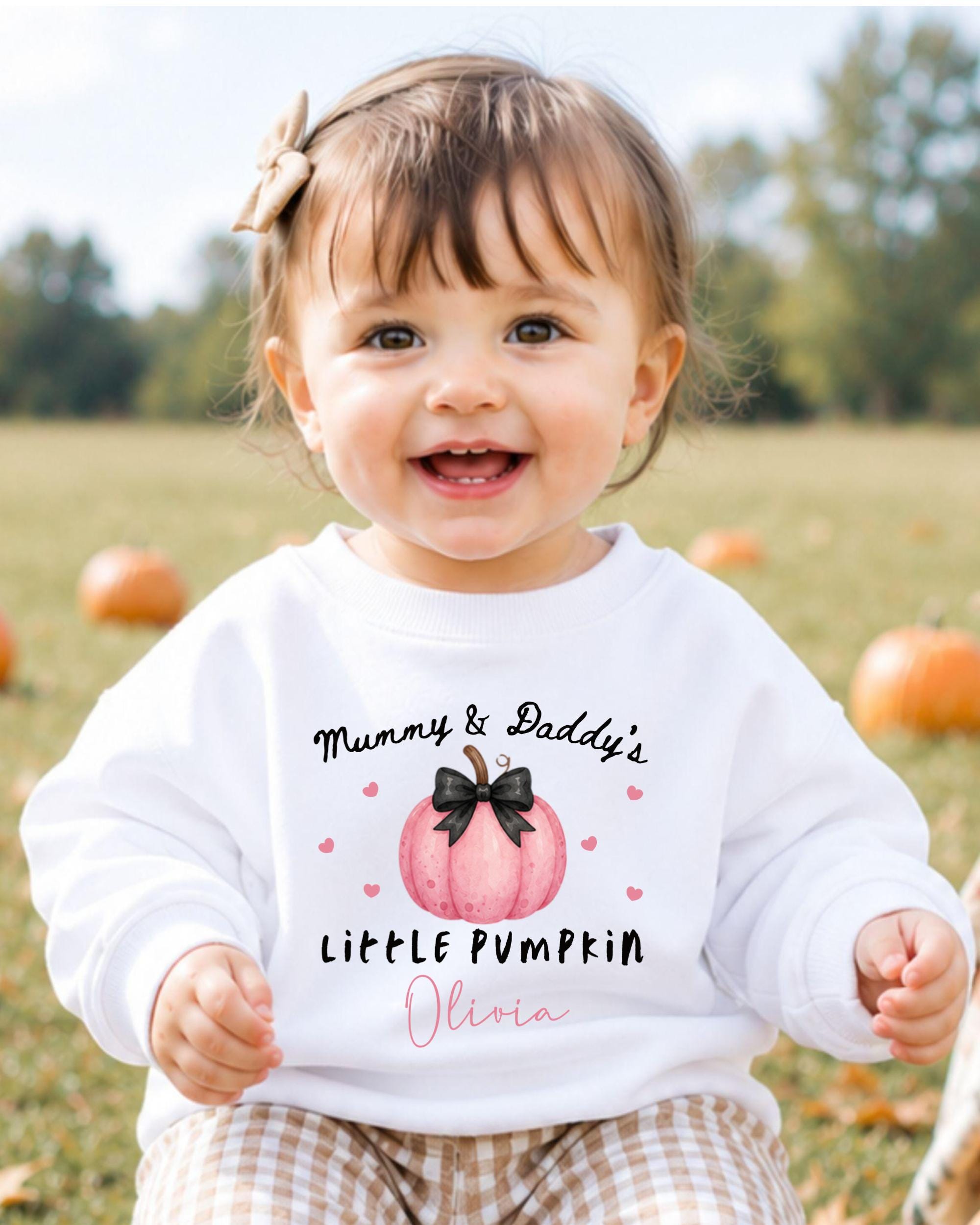 A smiling baby girl wearing a white sweater with a pumpkin design and text that says "Mummy & Daddy's Little Pumpkin Olivia".