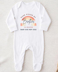 A white onesie with a graphic design on it, which says "Hand picked by Brother in Heaven Baby due Nov 2026".