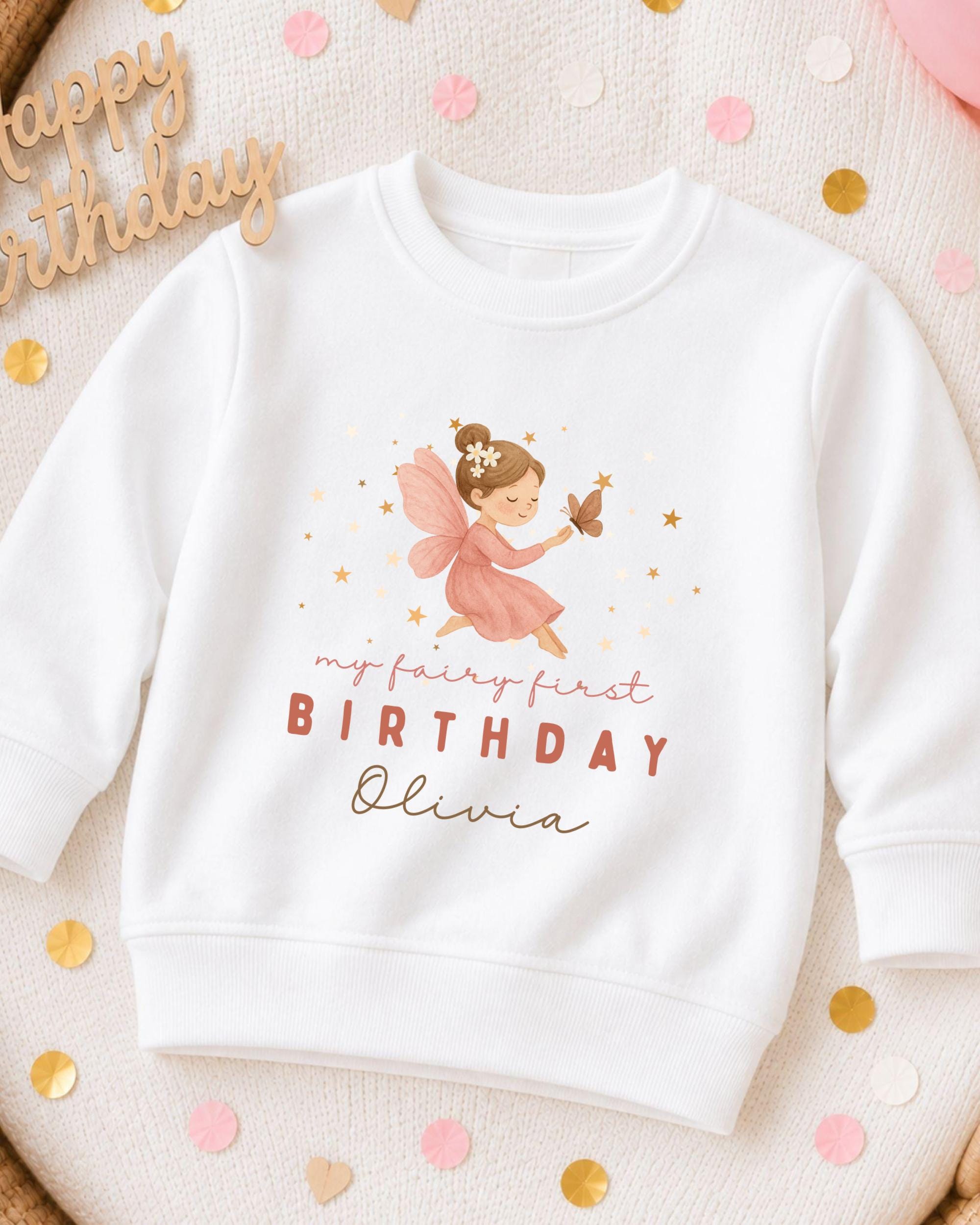 A white sweatshirt with a graphic of a fairy and the text "My fairy first birthday Olivia" printed on it, along with a "Happy birthday" sign.