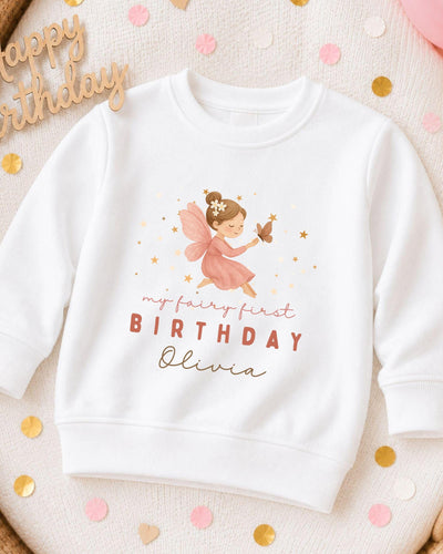 A white sweatshirt with a graphic of a fairy and the text "My fairy first birthday Olivia" printed on it, along with a "Happy birthday" sign.