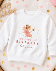 A white sweatshirt with a graphic of a fairy and the text "My fairy first birthday Olivia" printed on it, along with a "Happy birthday" sign.