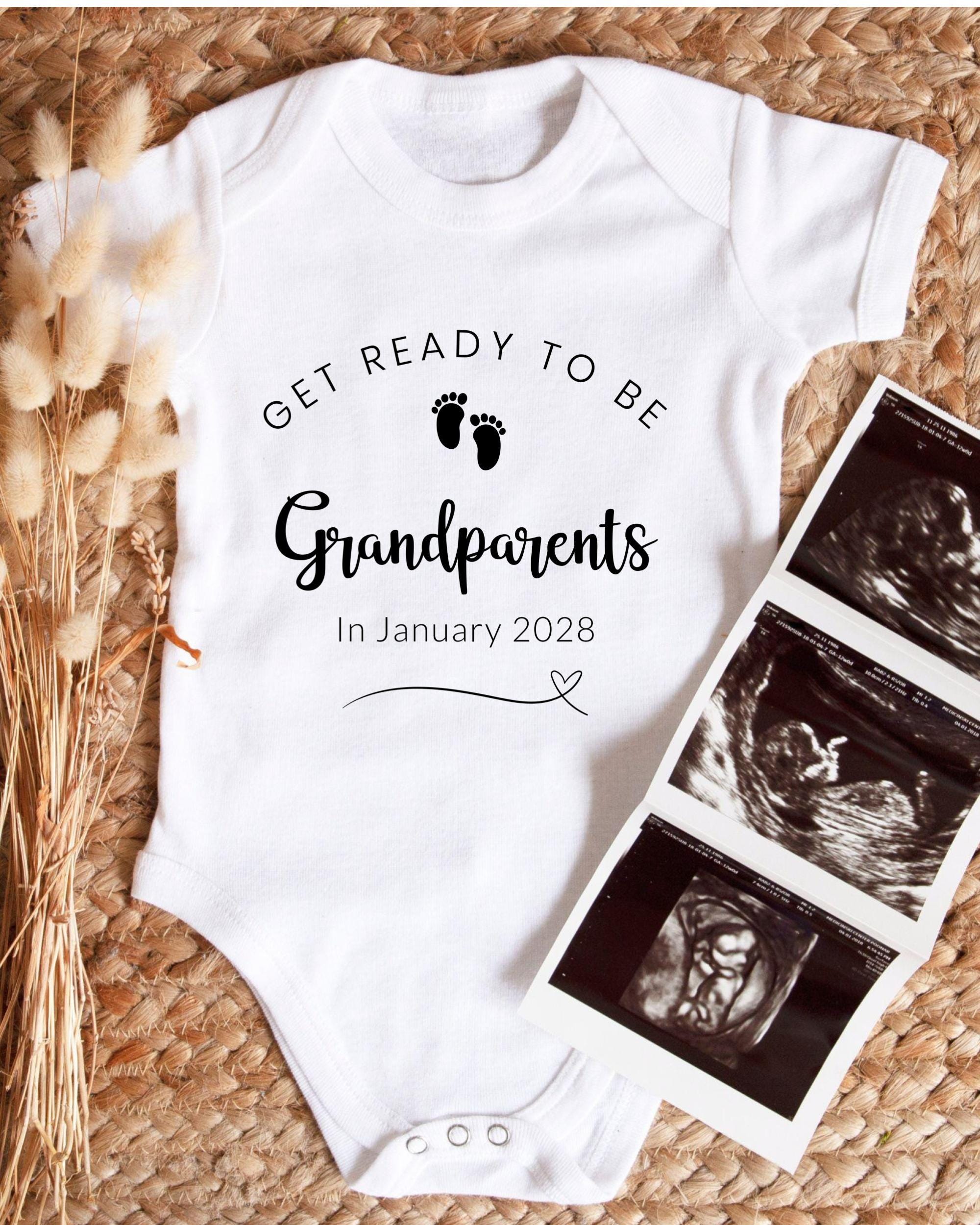 A white baby onesie with the text "GET READY TO BE Grandparents In January 2028" printed on it, along with a pair of black and white baby footprints. The onesie is placed next to a set of three black and white baby ultrasound images.