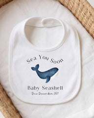 A white bib with a graphic of a blue whale and text that reads "Sea You Soon Baby Seashell".