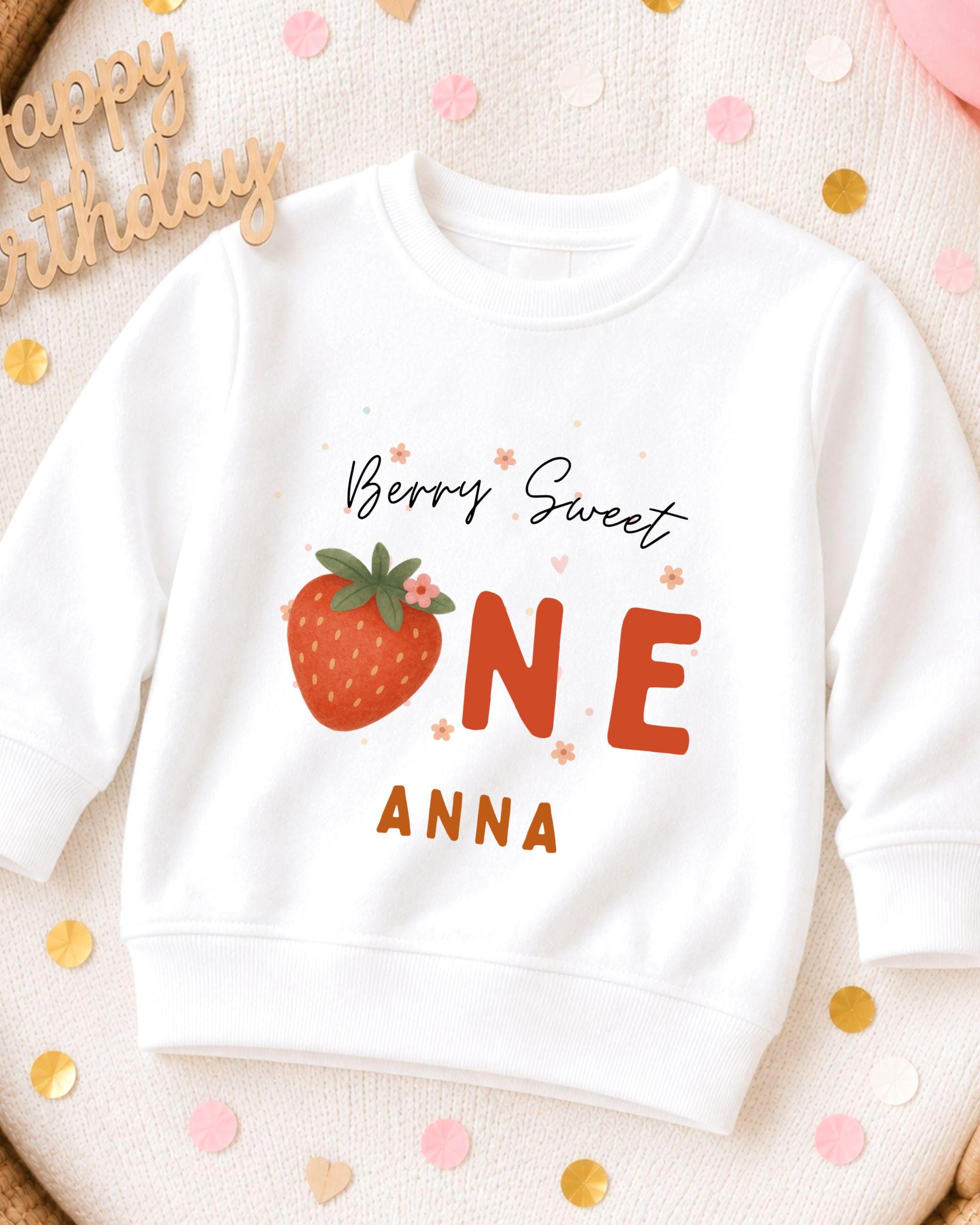 A white sweatshirt with a strawberry design and text printed on it, placed on a surface with confetti.