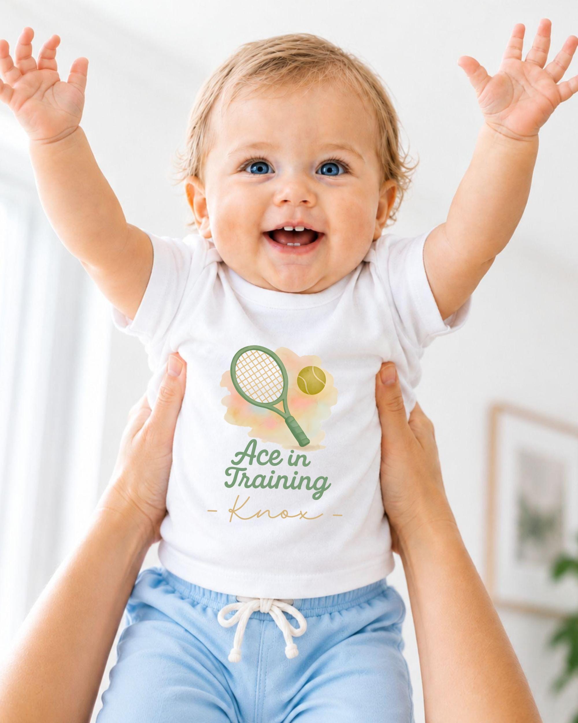 A smiling baby wearing a white shirt with a tennis racket and ball graphic, being held up by someone's arms.