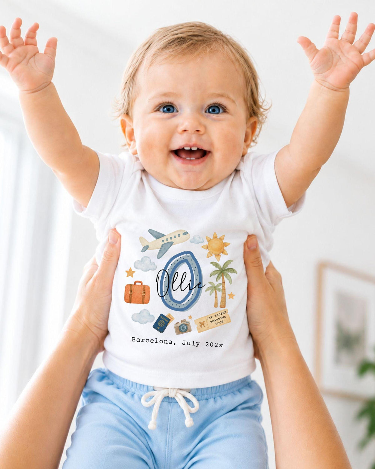 A smiling baby wearing a white shirt with a graphic design and text, being held up by someone's arms.