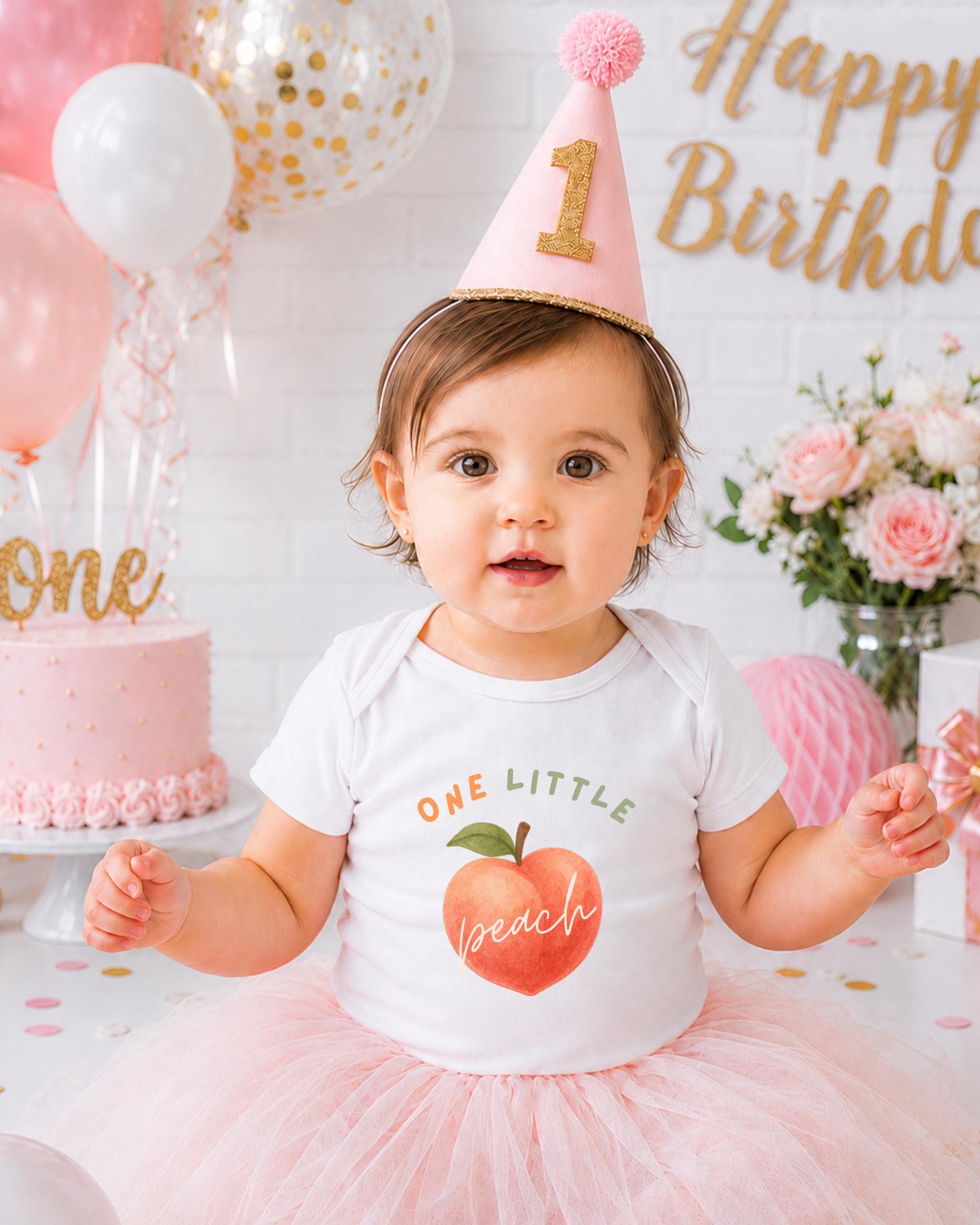 A young girl wearing a white t-shirt with a peach graphic and a pink birthday hat sits in front of a pink and white birthday cake, surrounded by balloons and flowers.