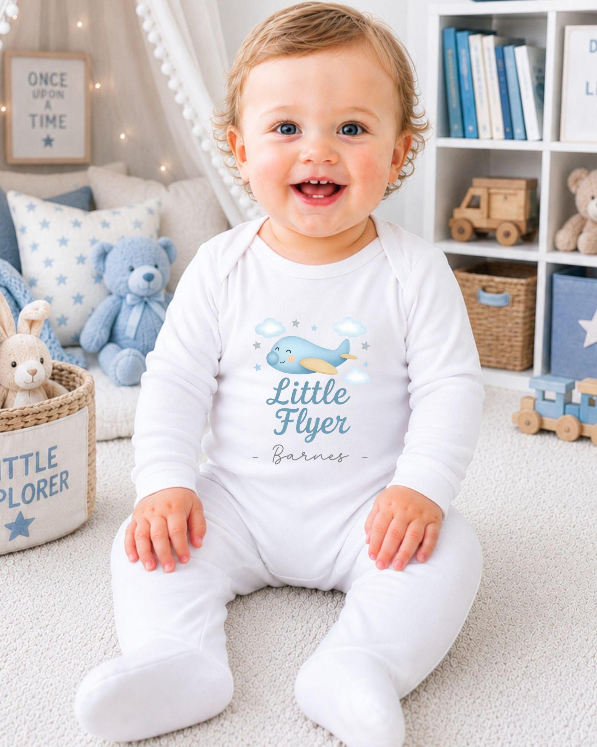 A smiling baby boy wearing a white onesie with a blue airplane design and the text "Little Flyer" is sitting on the floor in a nursery setting.