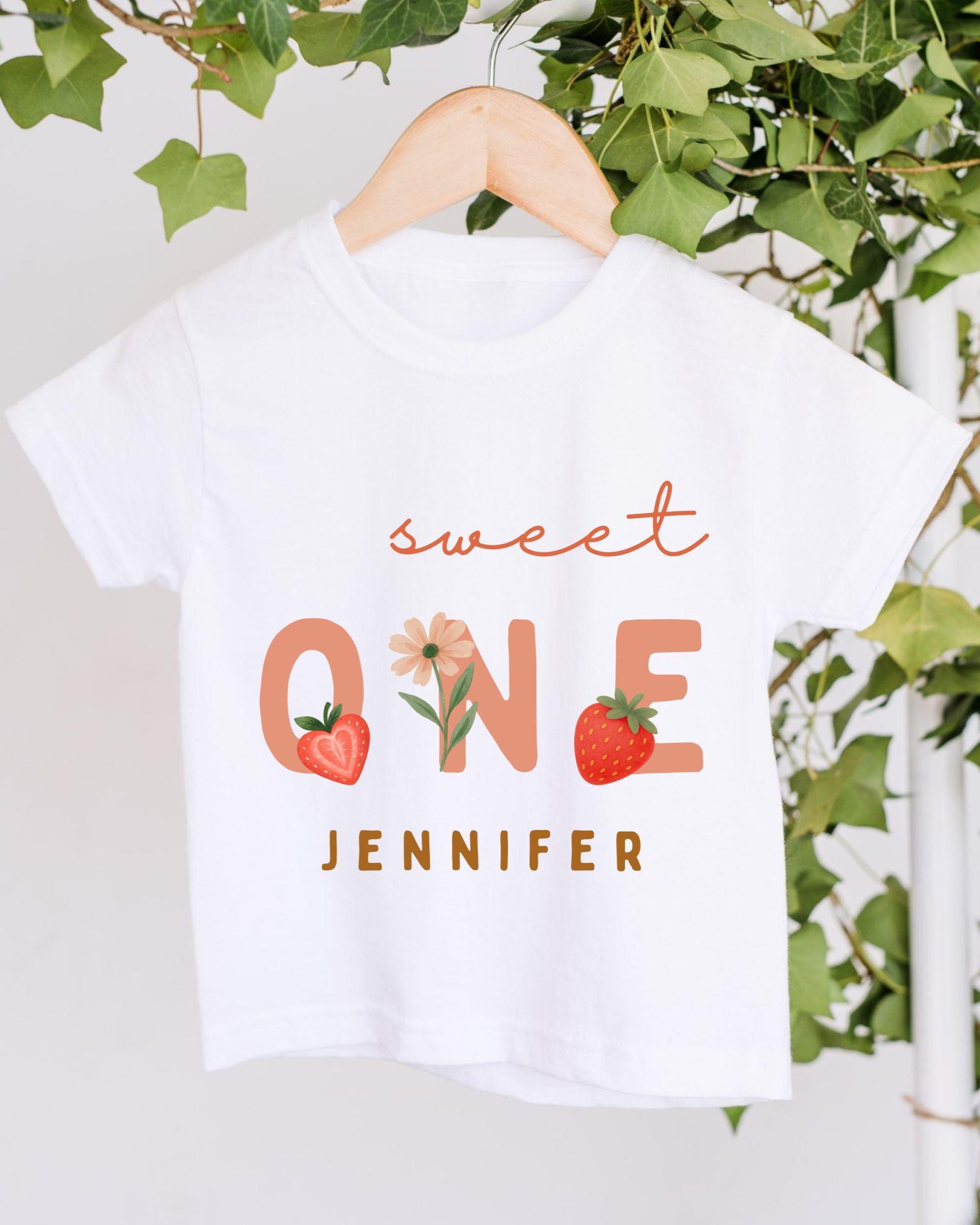 A white t-shirt with the text "Sweet One" and "JENNIFER" printed on it, along with illustrations of a strawberry and a flower.