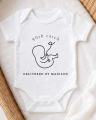 A white onesie with a graphic of a baby and text that reads "40th catch delivered by Madison".