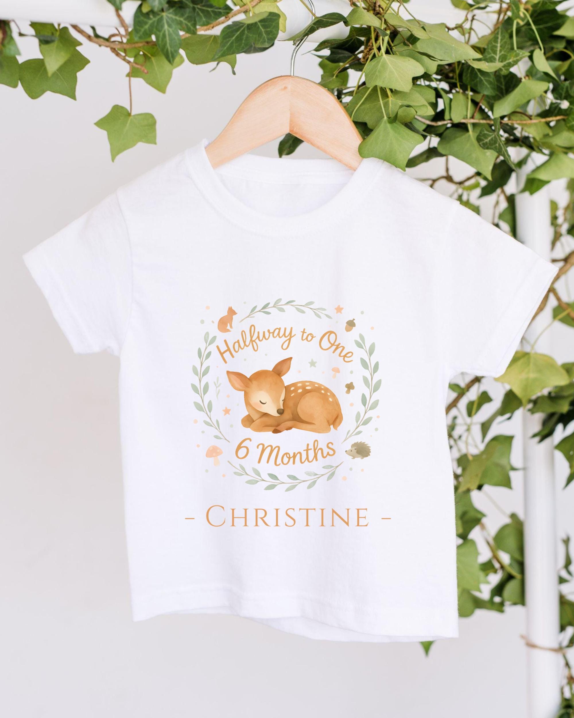 A white t-shirt with a graphic design featuring a baby deer and the text "Halfway to One 6 Months Christine" hanging on a wooden hanger.