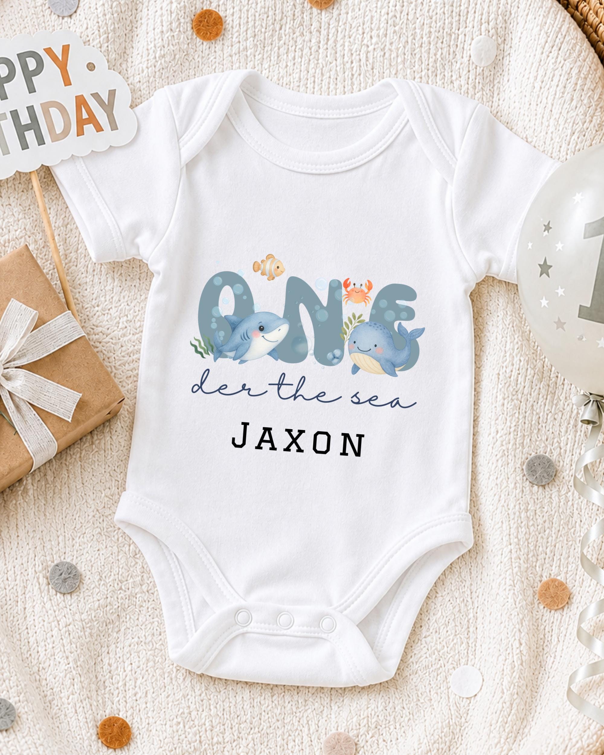 A white onesie with a design of a shark and other sea creatures, along with the text "One der the sea JAXON" printed on it, is displayed on a bed with a "Happy Birthday" sign and confetti.