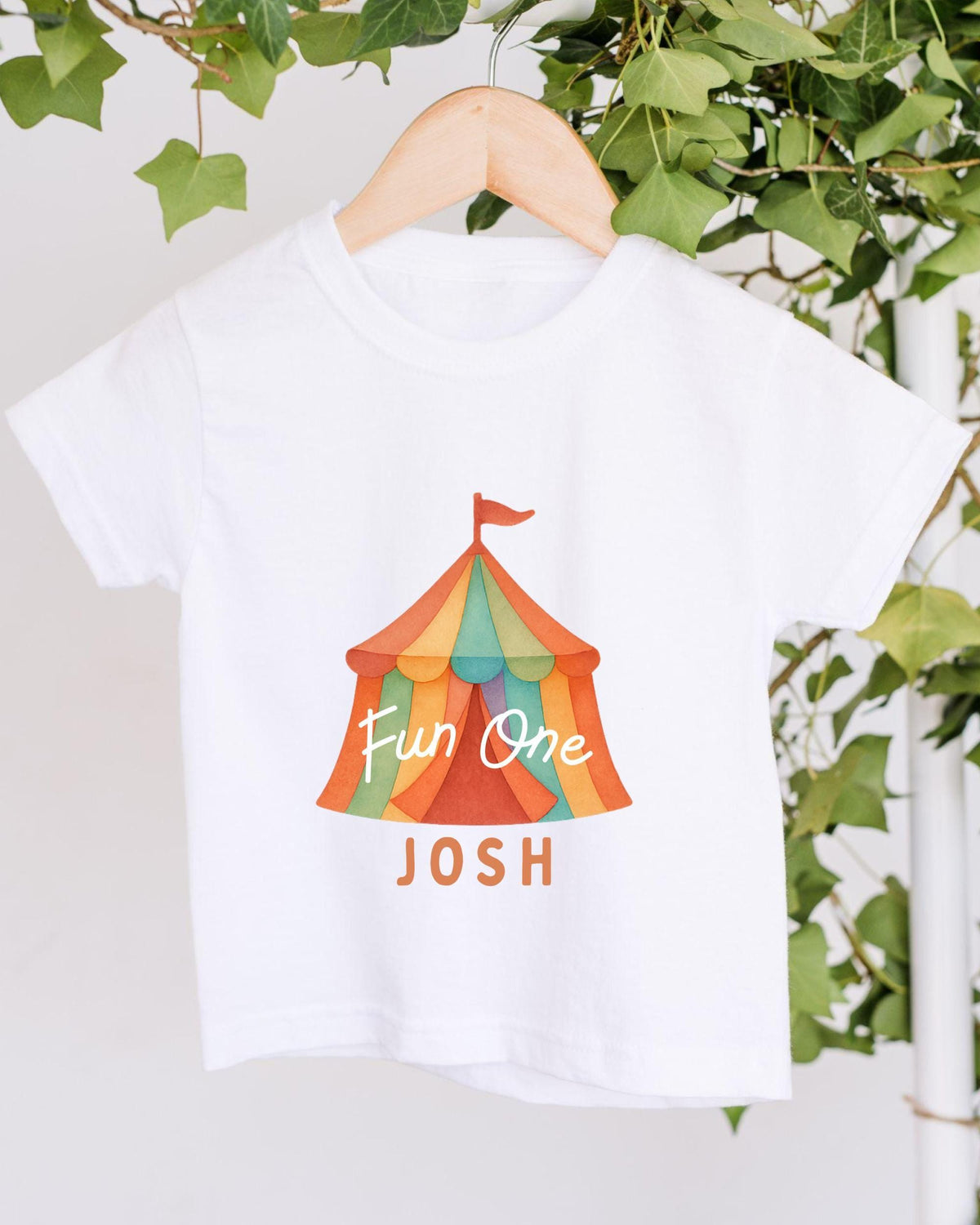 A white t-shirt with a colorful circus tent graphic and the text "Fun One Josh" printed on it, hanging on a wooden hanger.