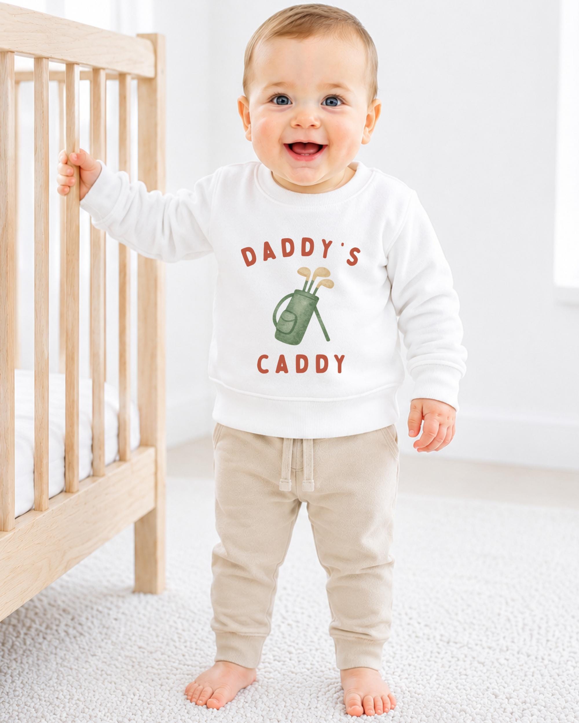A smiling baby boy wearing a white sweatshirt with the text "DADRY'S CADDY" printed on it, standing next to a wooden crib.