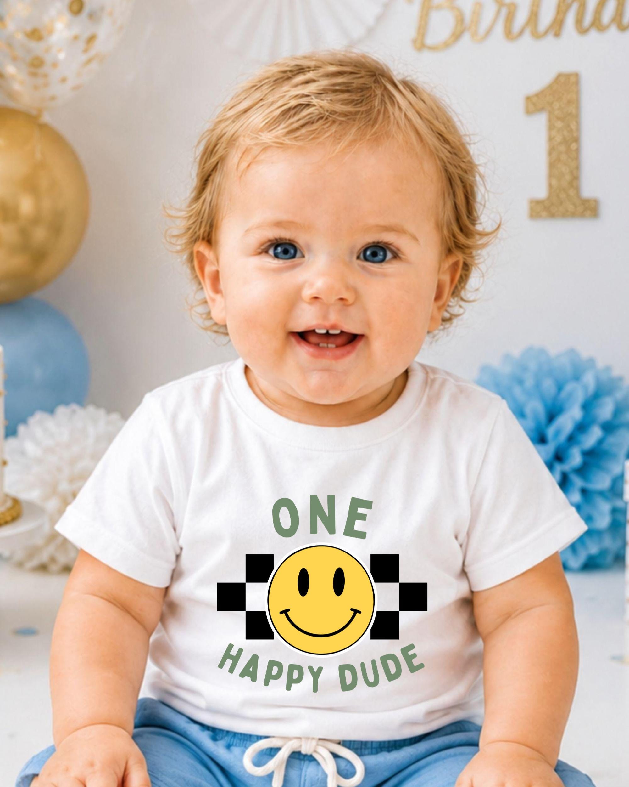 A smiling baby boy wearing a white t-shirt with a happy smiley face and the text "One Happy Dude" is sitting in front of a birthday party backdrop.