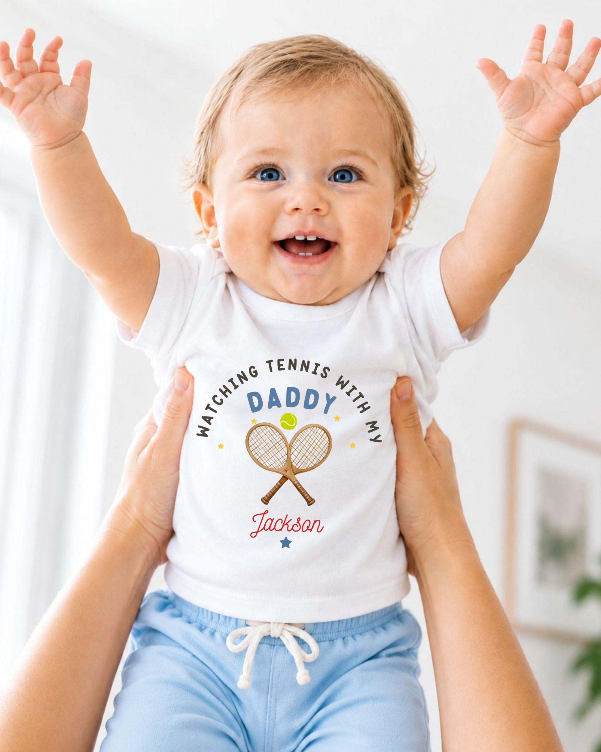 A smiling baby wearing a white shirt with a tennis racket and the text "Watching Tennis with my Daddy Jackson" is being held up by someone's arms.