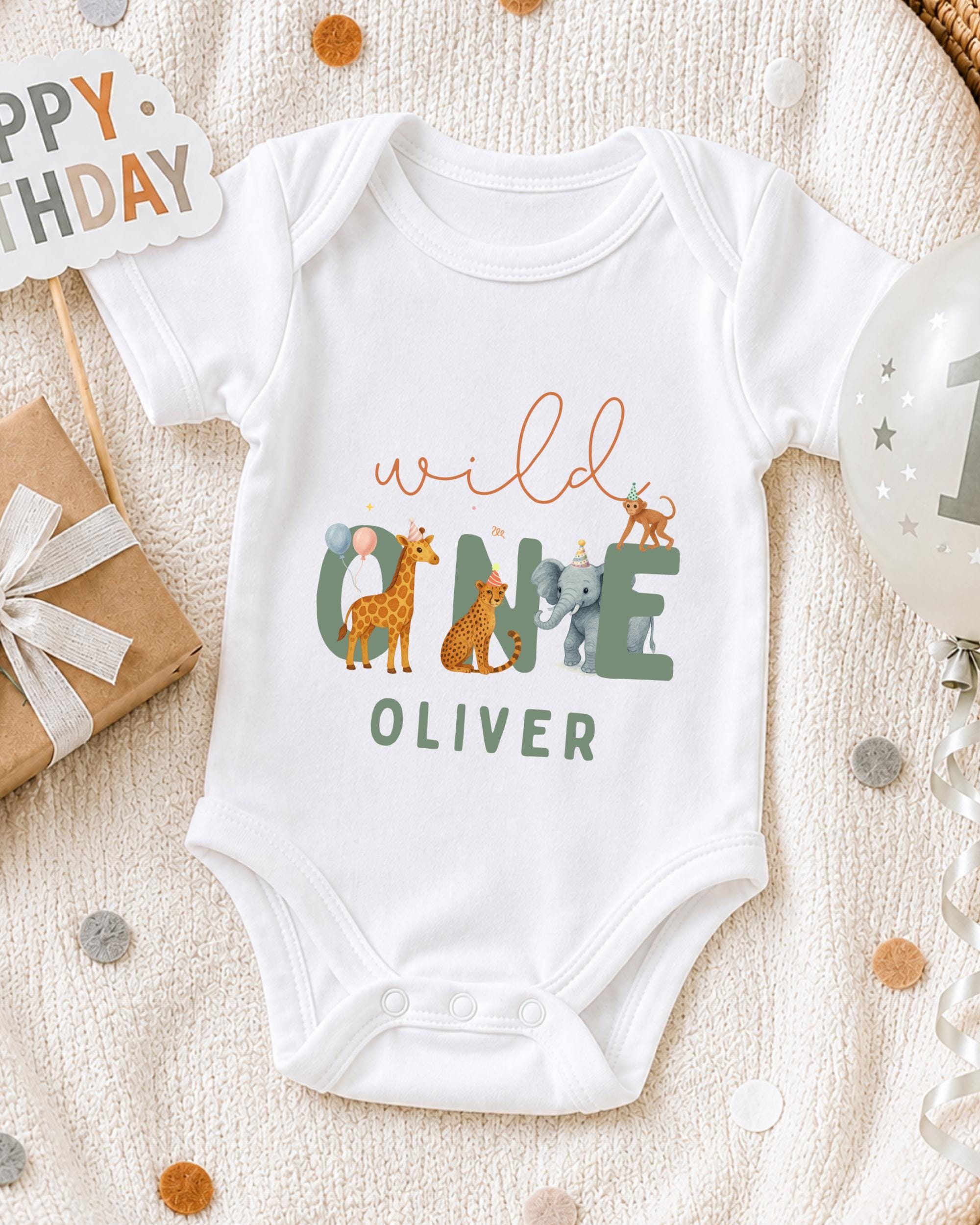 A white onesie with the text "wild Oliver" printed on it, surrounded by various animal illustrations, is displayed on a bed with a "Happy Birthday" sign and confetti.