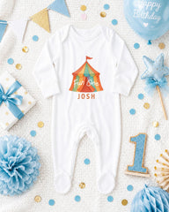 A baby onesie with a circus tent design, surrounded by birthday decorations such as a blue balloon, blue and gold confetti, and a blue and gold bow.