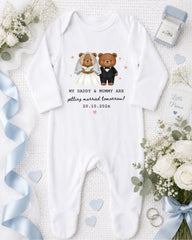 A white onesie with a graphic of a bride and groom, along with the text "My Daddy & Mummy are getting married tomorrow! 20.10.2021", is displayed on a bed with white flowers and a blue ribbon.