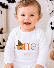 A smiling baby boy wearing a white onesie with a pumpkin and witch hat, sitting in front of Halloween decorations.