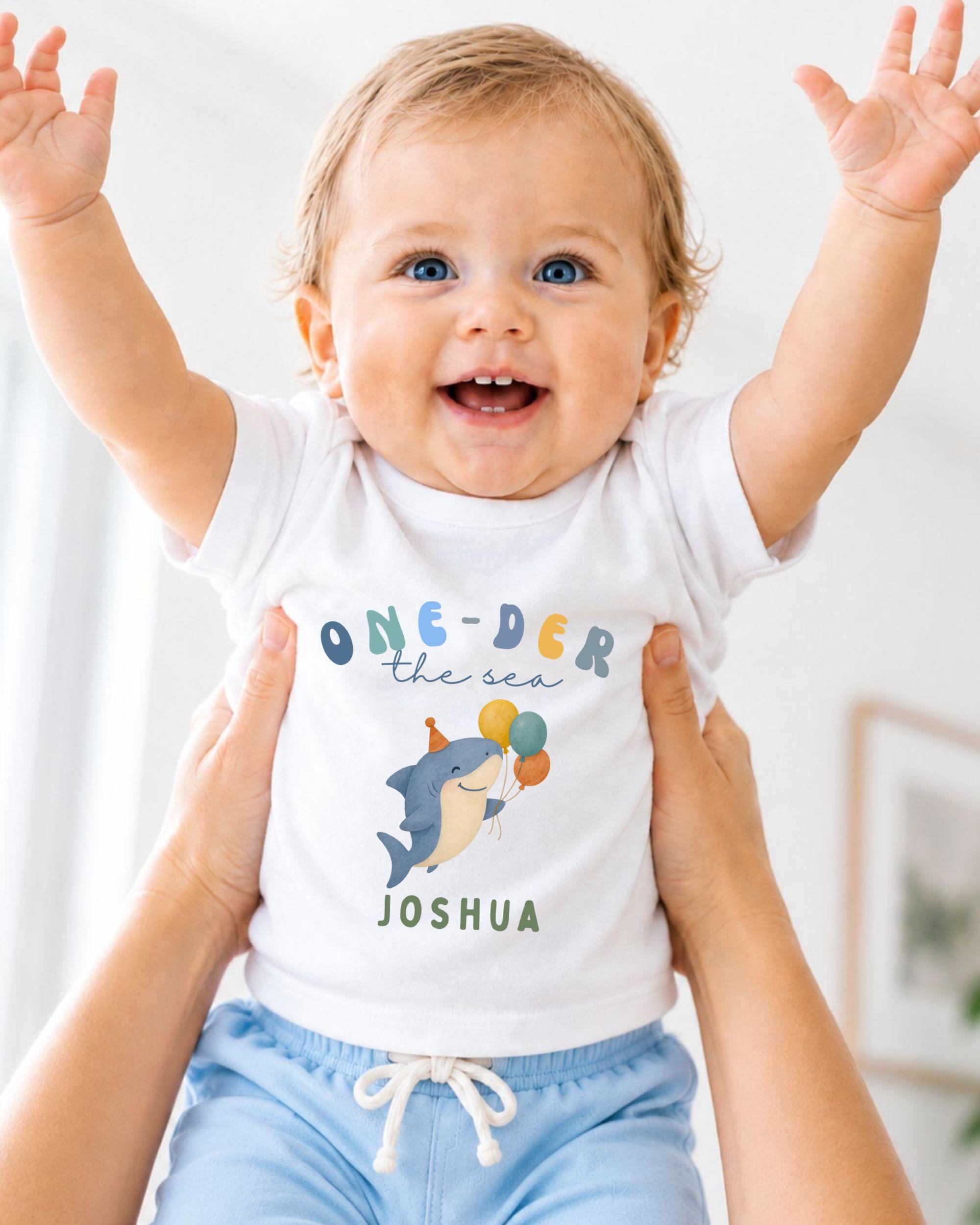 A smiling baby wearing a white t-shirt with a shark graphic and the text "One-DER the sea Joshua" is being held up by someone's arms.