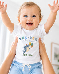 A smiling baby wearing a white t-shirt with a shark graphic and the text "One-DER the sea Joshua" is being held up by someone's arms.