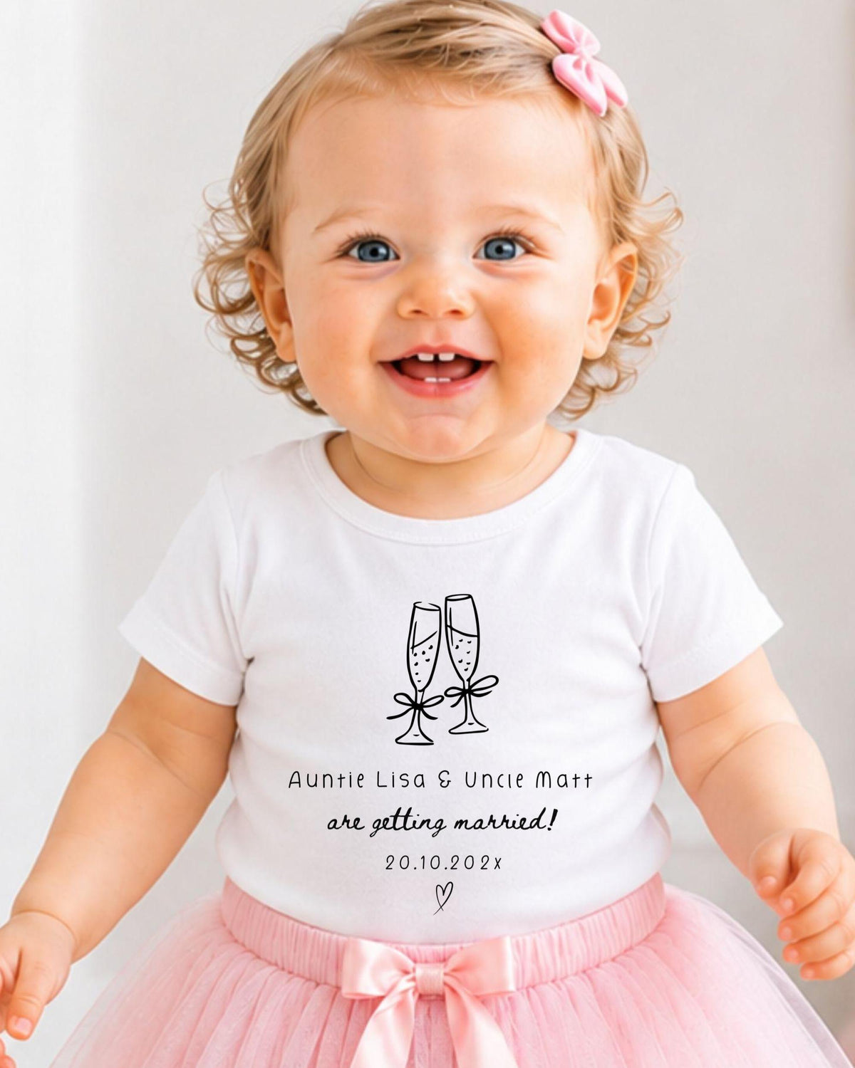 A smiling baby girl wearing a white t-shirt with a graphic and text, standing in front of a white background.