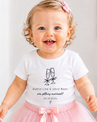 A smiling baby girl wearing a white t-shirt with a graphic and text, standing in front of a white background.