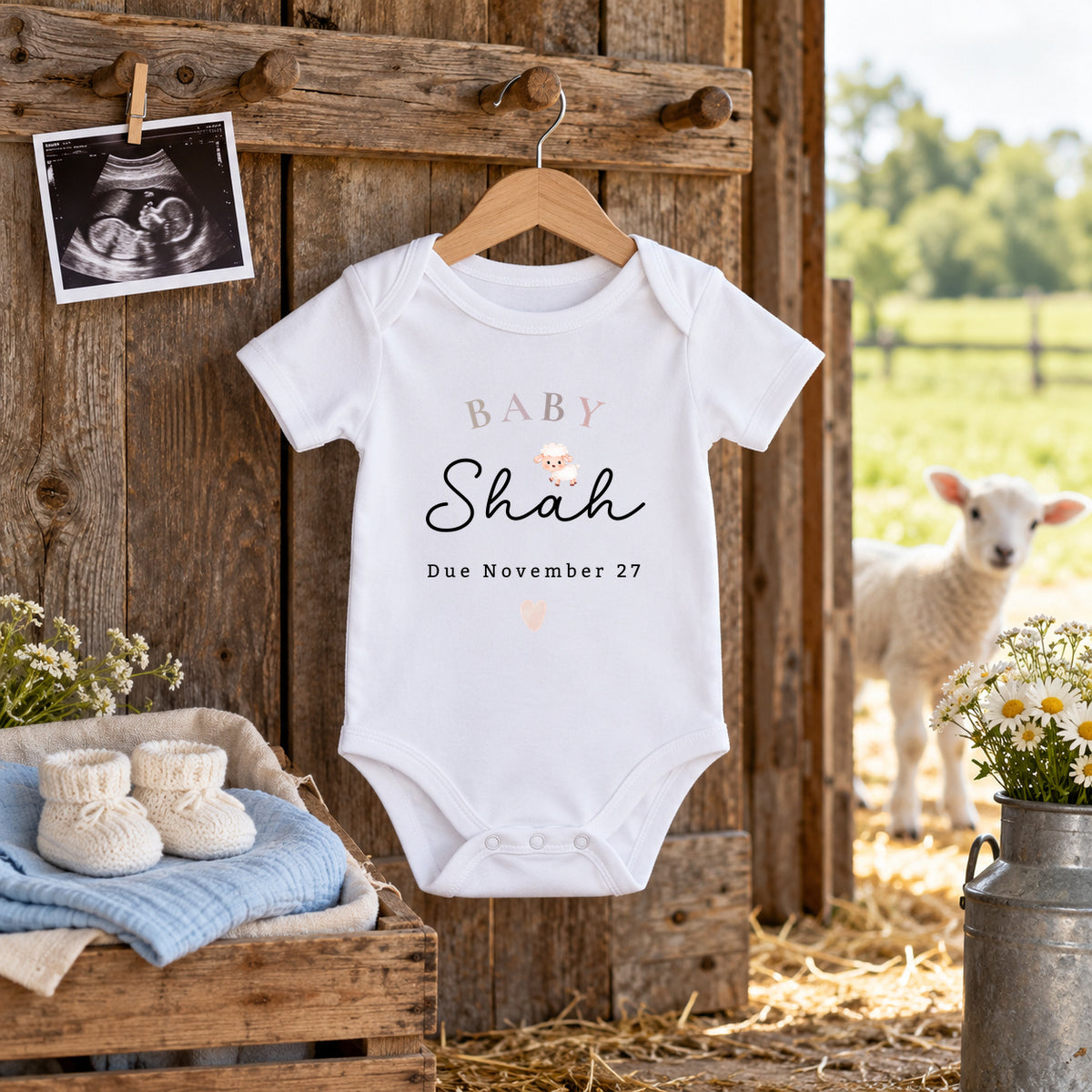 White Baby Shah lamb Baby Vest reading Due November 27, hanging in a barn doorway beside a scan photo, cream booties on blue muslin, daisies in a milk churn and a lamb outside.
