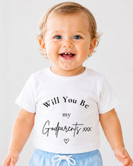 A smiling baby wearing a white t-shirt with text that says "Will You Be my Godparents xxx" printed on it.